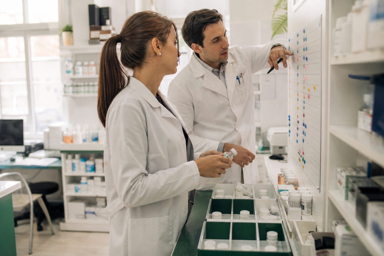Two pharmacists reviewing a wall planner of colored dot stickers at the back counter of a community pharmacy